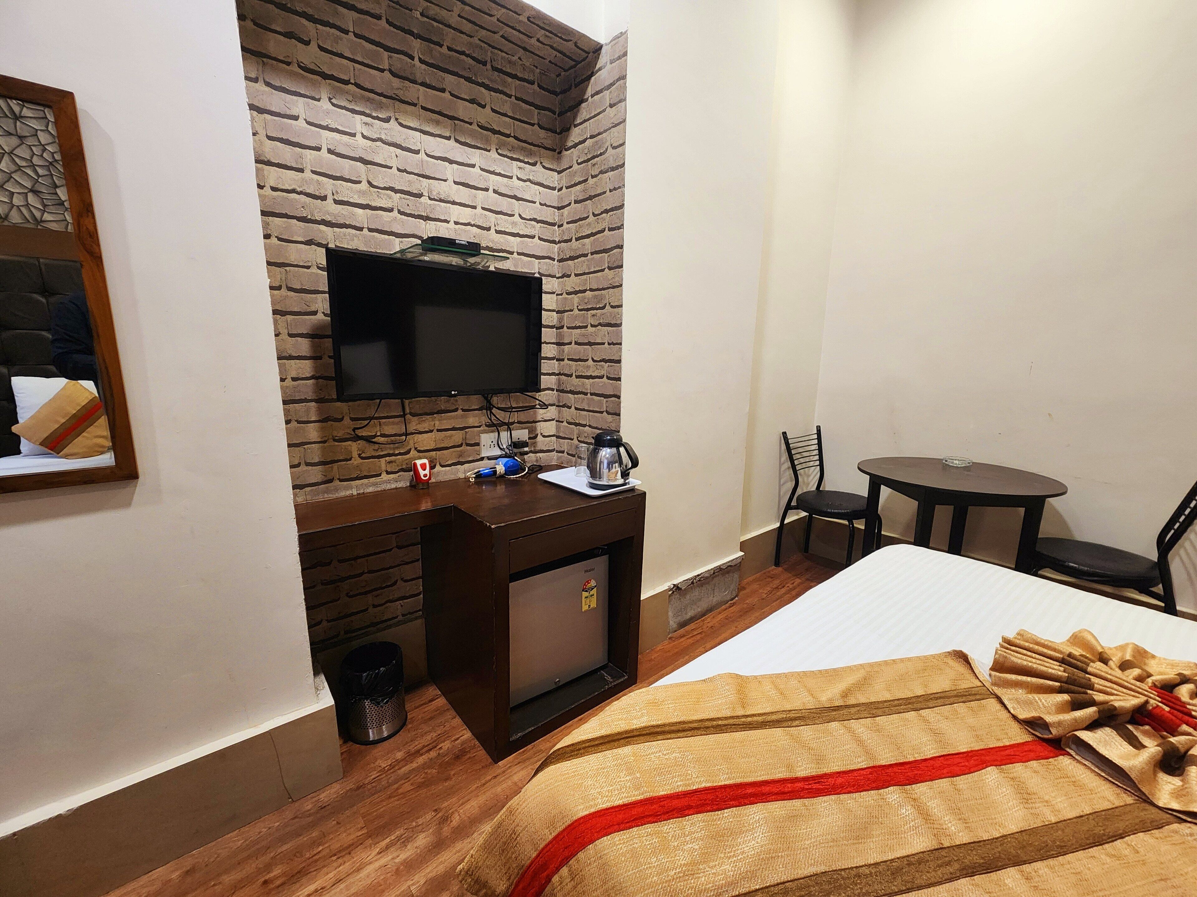 A hotel room with a brick wall, a flat-screen TV mounted above a wooden desk, a small table with a chair, and a bed with a decorative blanket.