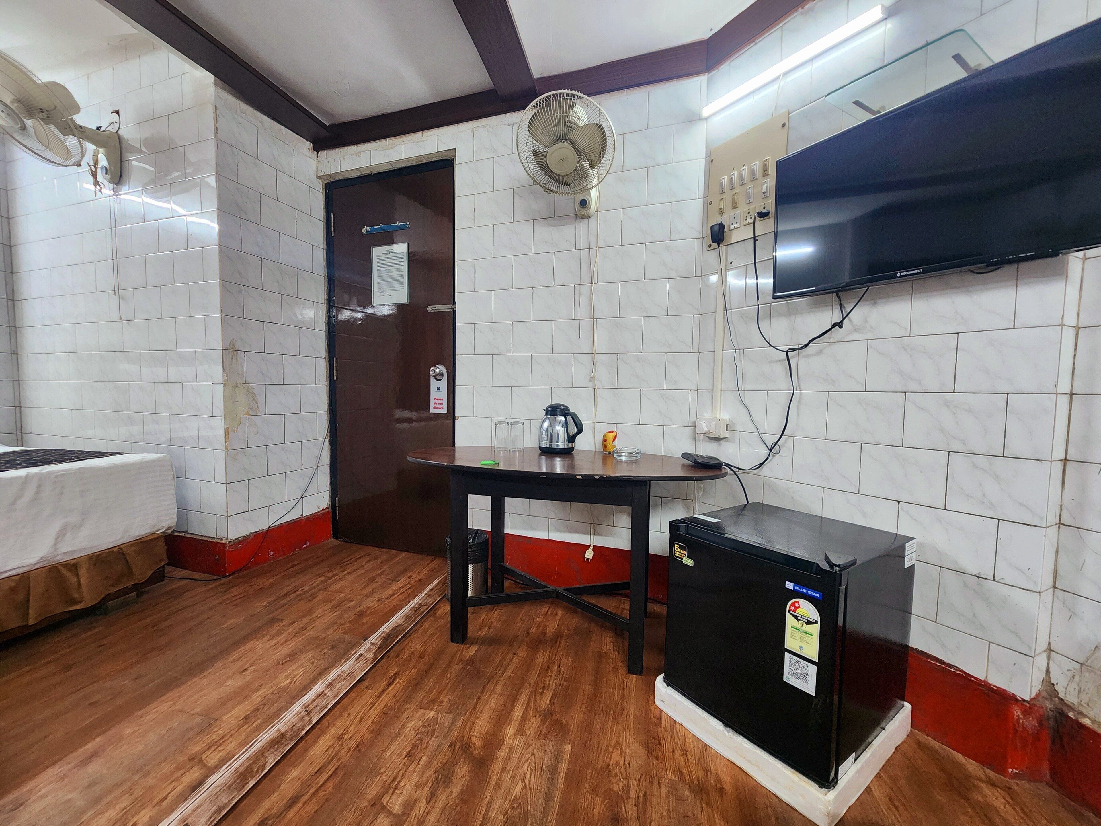 A room with a wooden floor, a table with a kettle and glasses, a black refrigerator, a TV mounted on the wall, and a door with a sign.