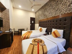 Hotelkamer met twee bedden, een bakstenen muur, een stenen muur, een plafondventilator en een bureau met koffiezetapparaat.