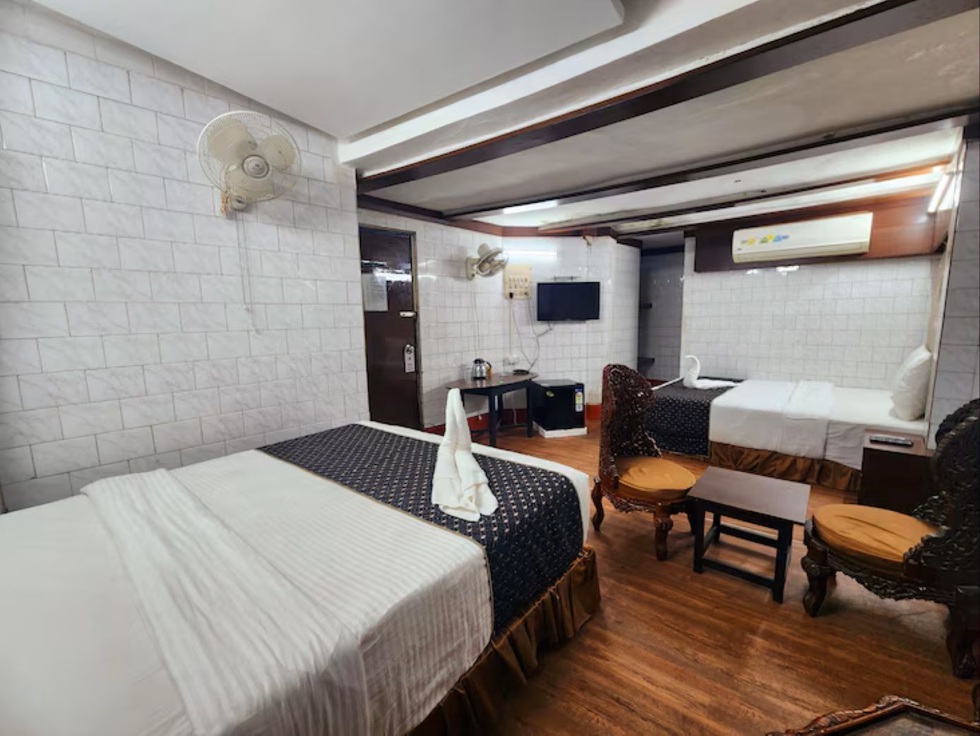 Ein Hotelzimmer mit zwei Betten, einem kleinen Tisch, einem Sessel, einem Schreibtisch, einem Ventilator, einem Fernseher und einem Spiegel.