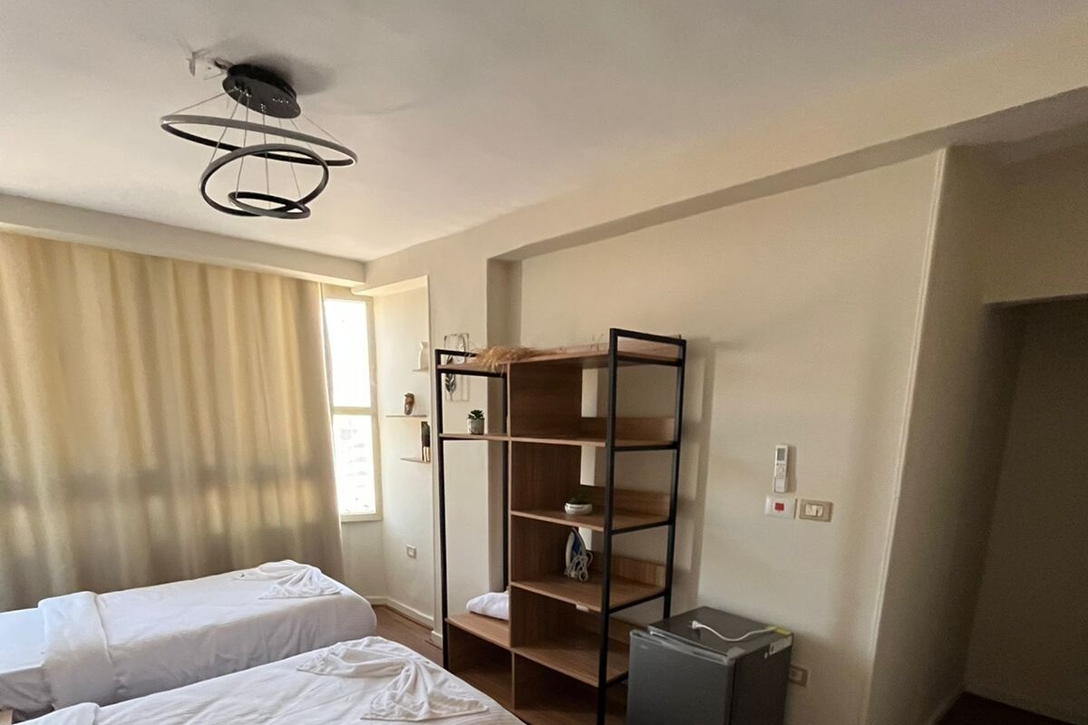 Habitación de hotel con dos camas individuales, un escritorio de madera, un refrigerador pequeño y una unidad de estanterías empotrada.