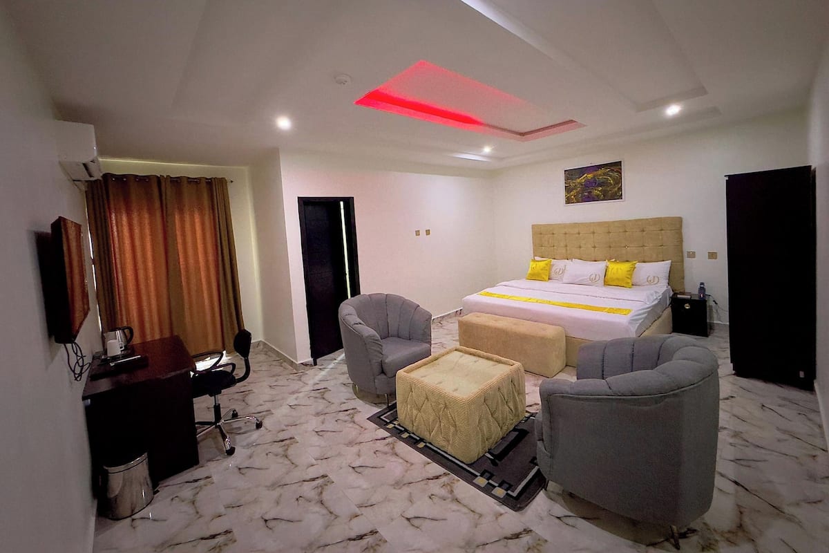 A hotel room with a bed, two armchairs, a small table, a desk with a chair, a TV, and a ceiling light.