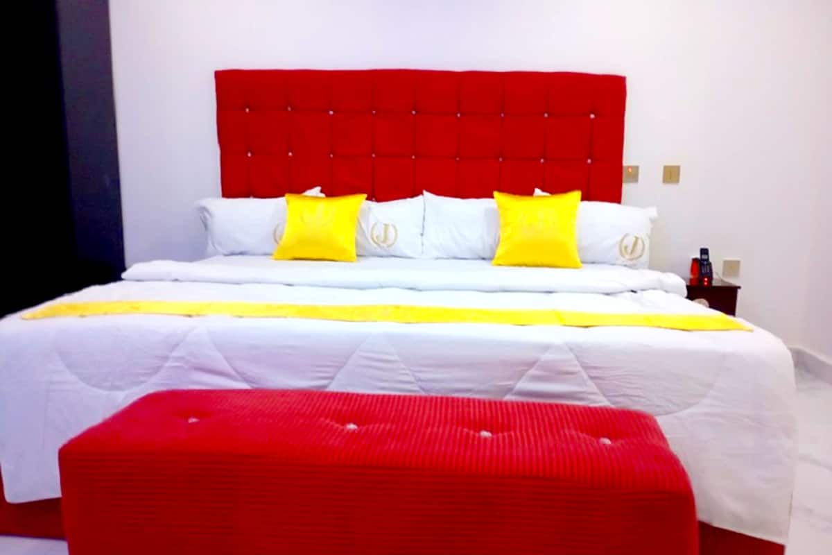 A hotel room with a large bed, red headboard, white bedding, and a red ottoman.