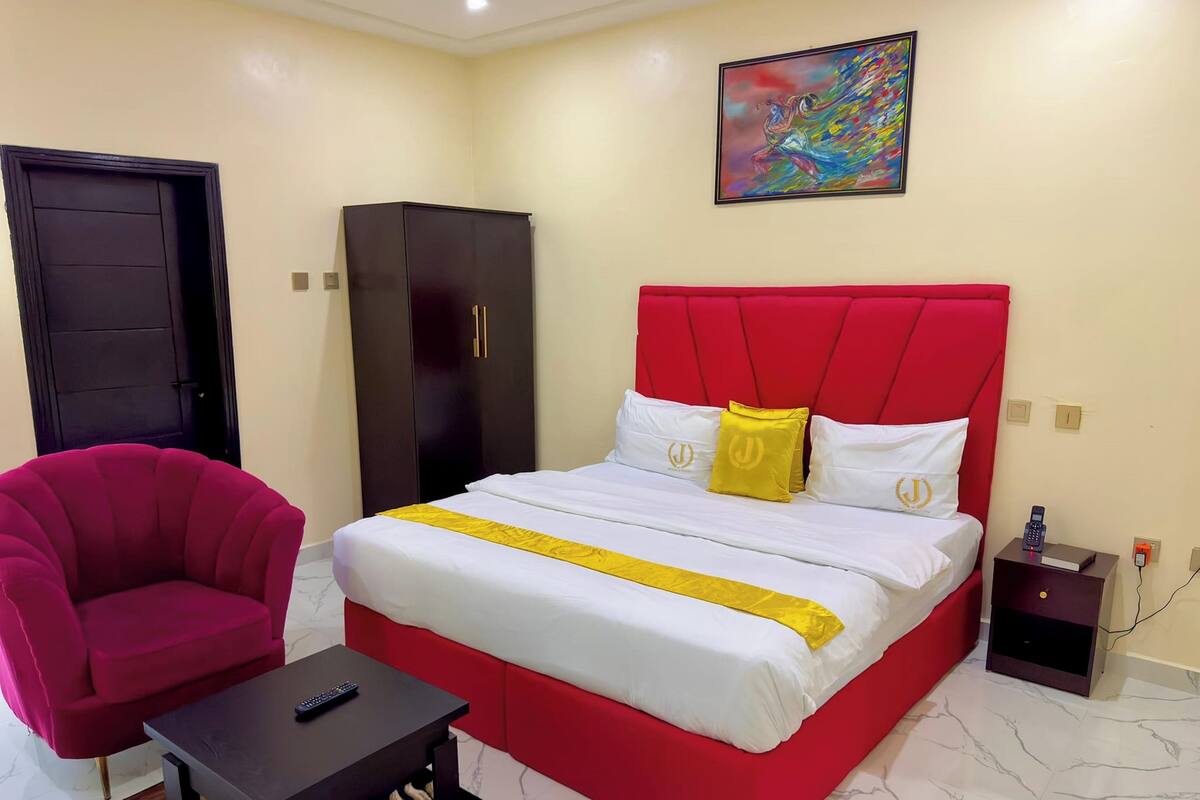A hotel room with a red bed, a black wardrobe, a red armchair, a small table with a remote control, and a painting on the wall.