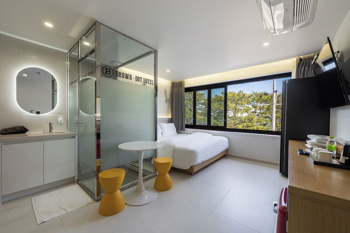 Habitación de hotel moderna con cabina de ducha de vidrio, cama con ropa de cama blanca, una mesita redonda y vistas a árboles a través de un ventanal grande.