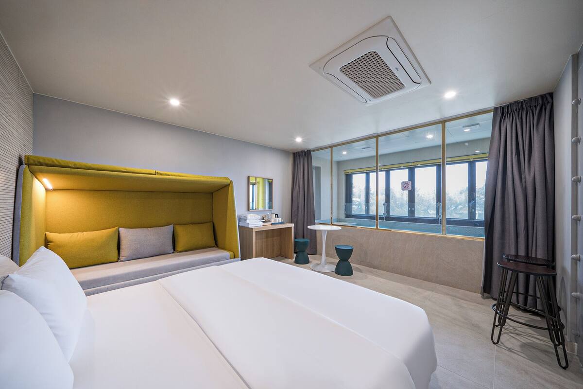 Habitación de hotel moderna con una cama grande, una mesita de noche, un taburete pequeño, una ventana con cortinas y vistas al exterior.