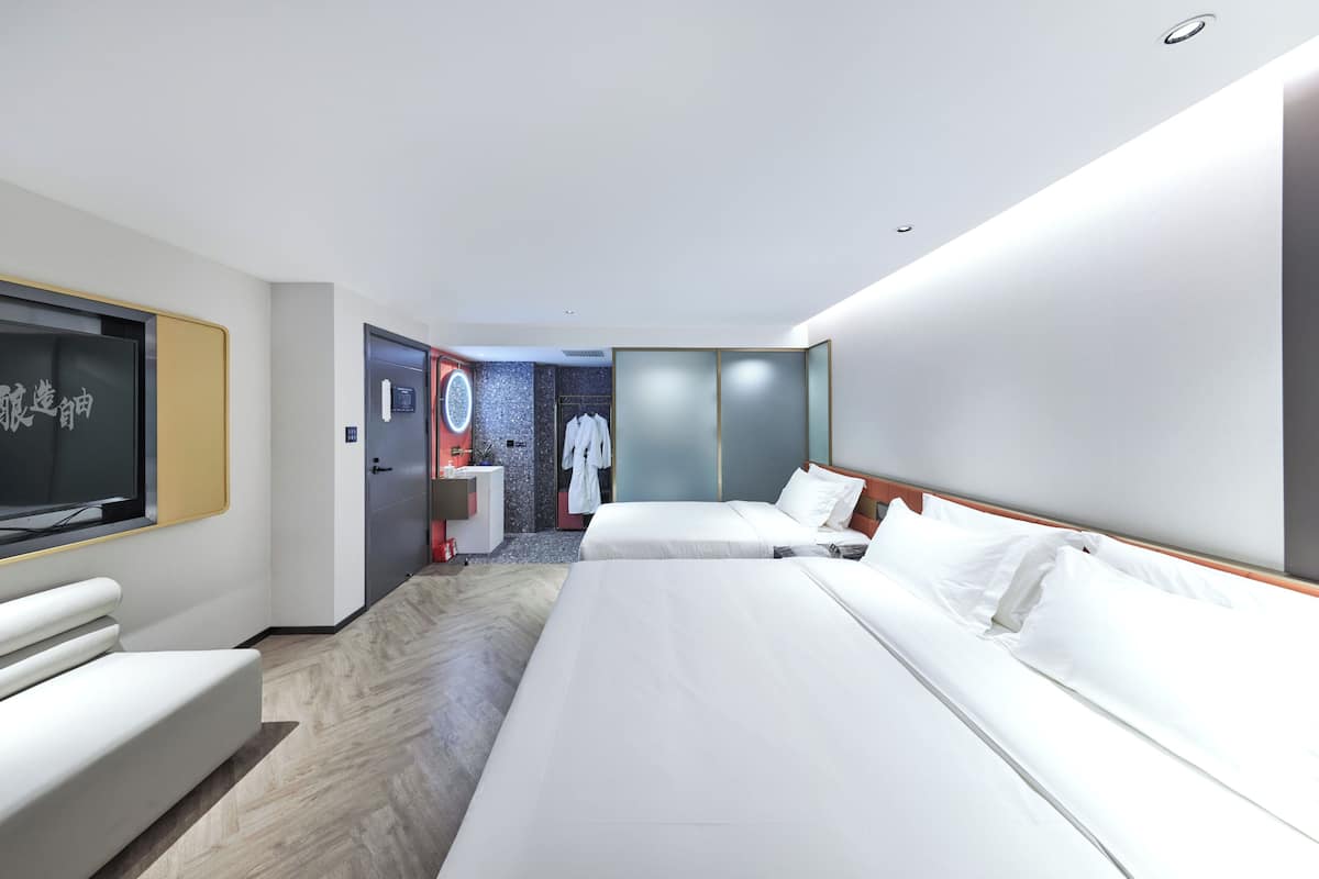 Une chambre d’hôtel moderne avec deux lits, un coin salon, une télévision et une salle de bain visible à travers une porte ouverte.