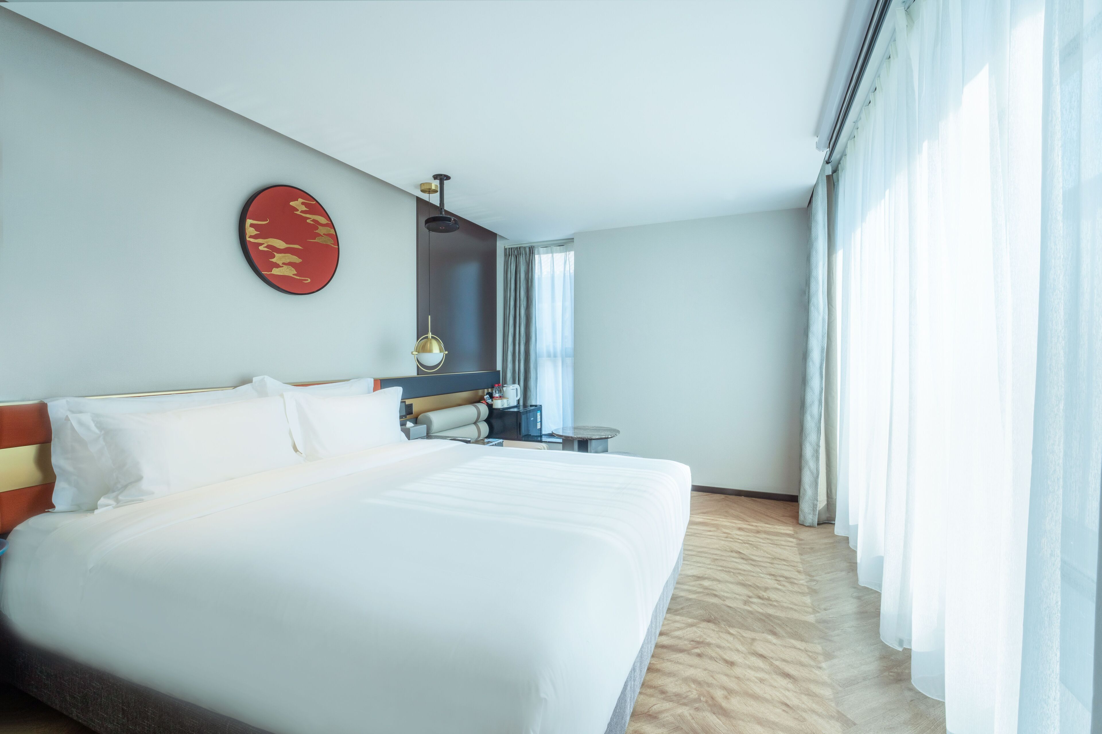 Habitación de hotel con una cama grande, una mesita de noche, un adorno rojo en la pared y una ventana con cortinas.
