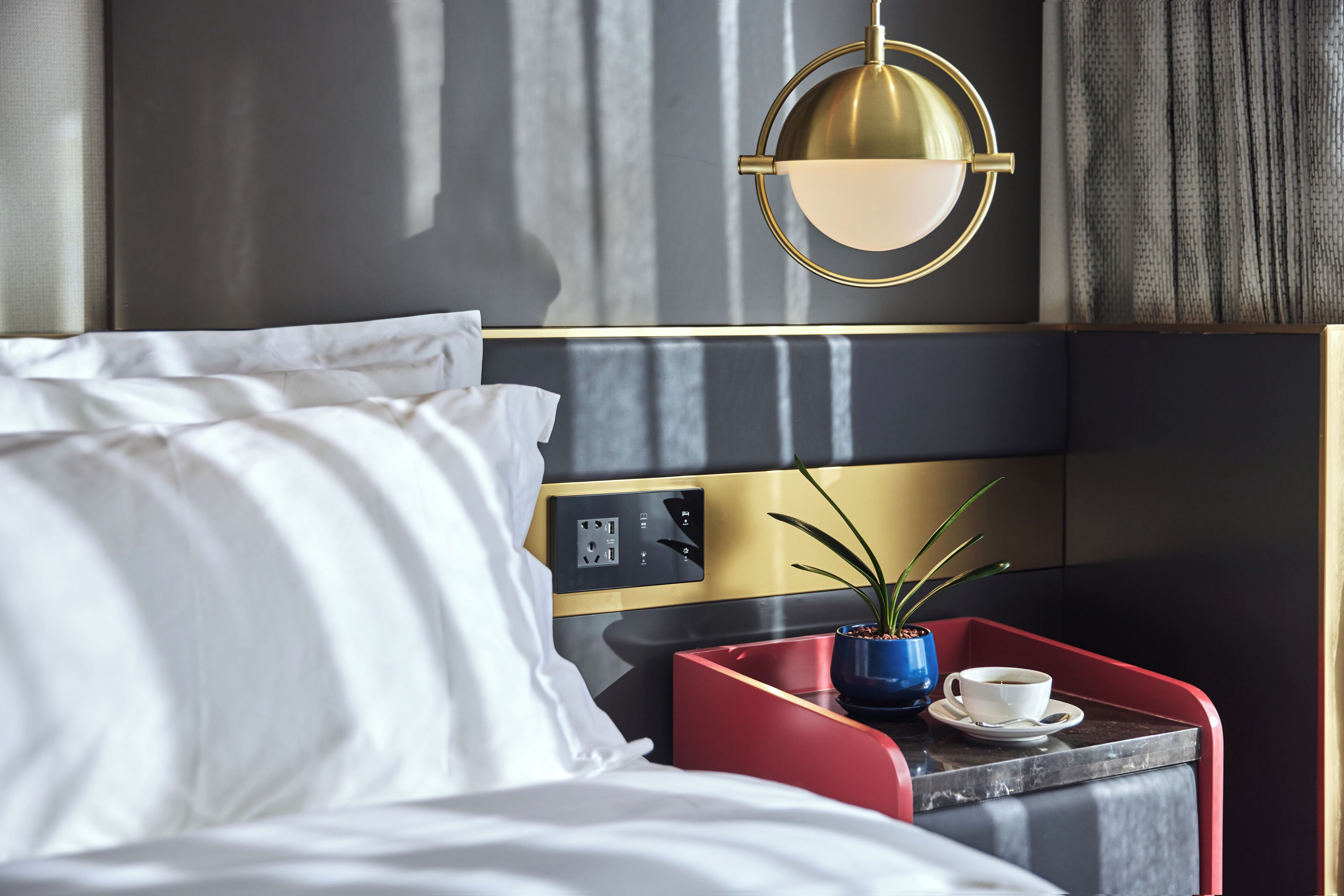 Una habitación de hotel moderna con una cama, una mesita de noche con una taza y un plato, una bandeja roja y un panel de control en la pared.