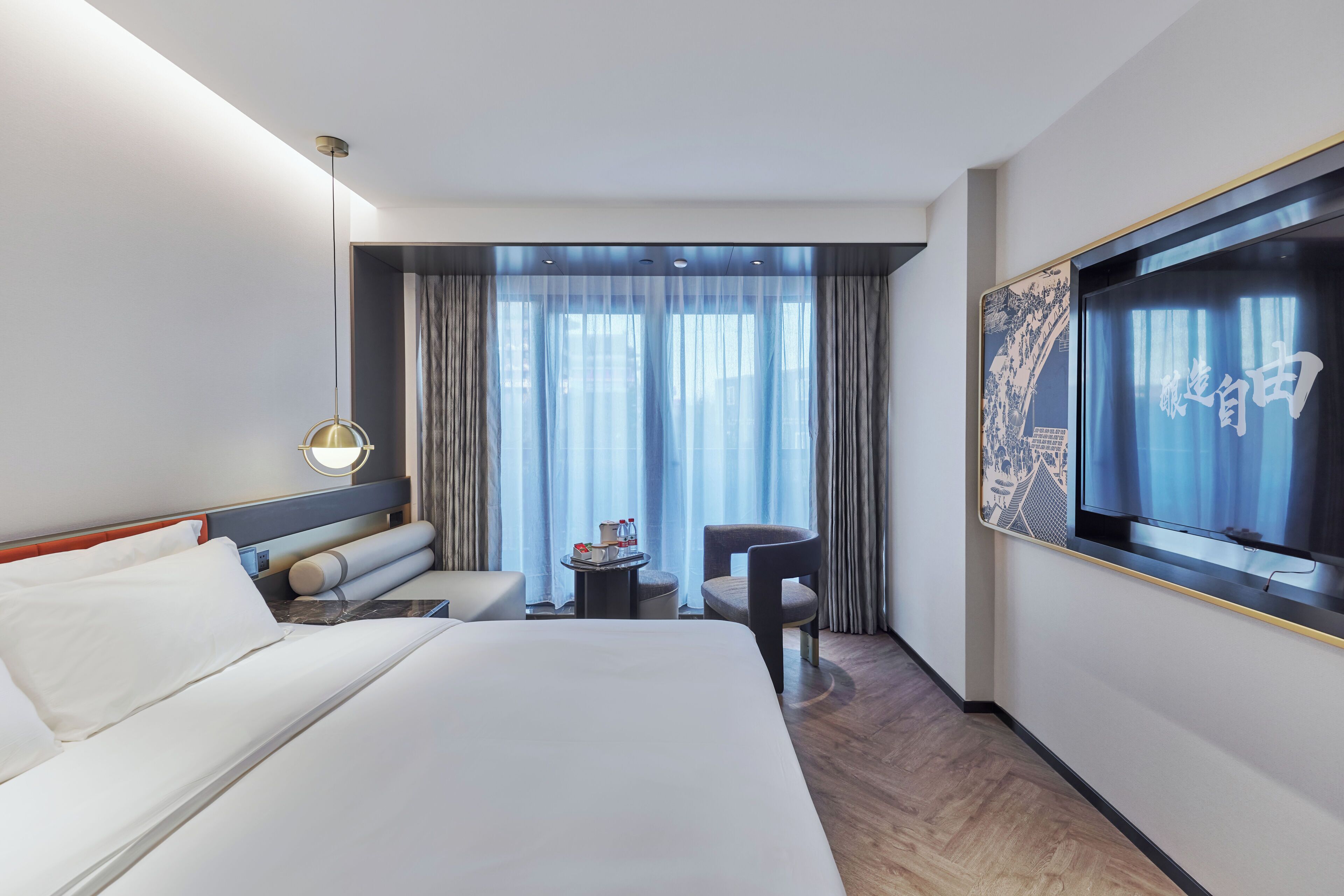 A hotel room with a large bed, a desk, a chair, a small table with a lamp, and a TV on the wall.
