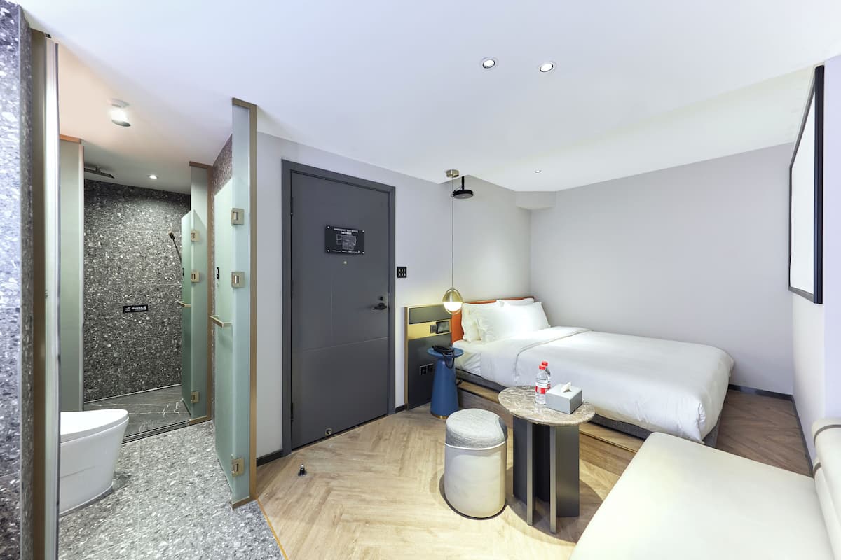 Une chambre d’hôtel avec un lit, une table de chevet et un petit tabouret. Il y a un espace douche avec une porte en verre et des toilettes.