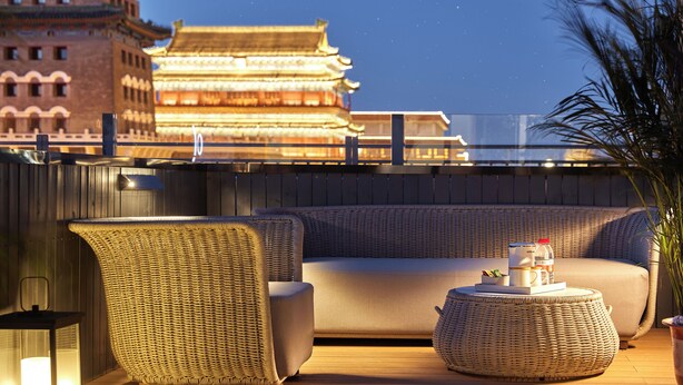 A rooftop lounge with a sofa, wicker chairs, and a round table. A traditional building with illuminated windows is visible in the background.