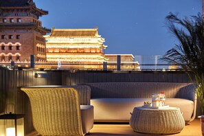 A rooftop lounge with a sofa, wicker chairs, and a round table. A traditional building with illuminated windows is visible in the background. - M Hotel  (Beijing)