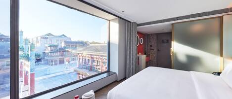A hotel room with a large window, a bed, a bedside table with a tray of items, and a view of traditional buildings outside.