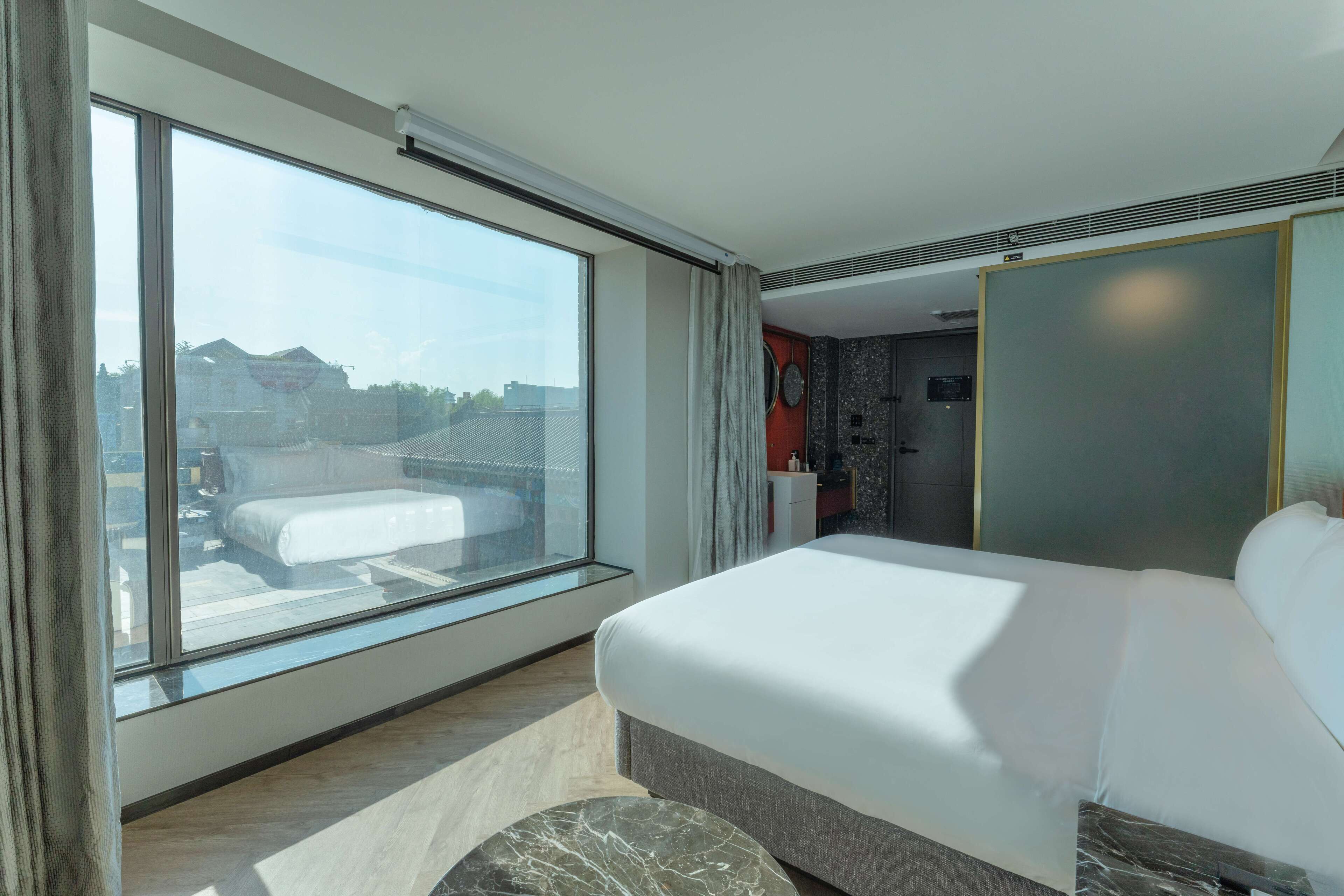 A modern hotel room with a large window, a bed, a marble accent piece, and a view of the cityscape.