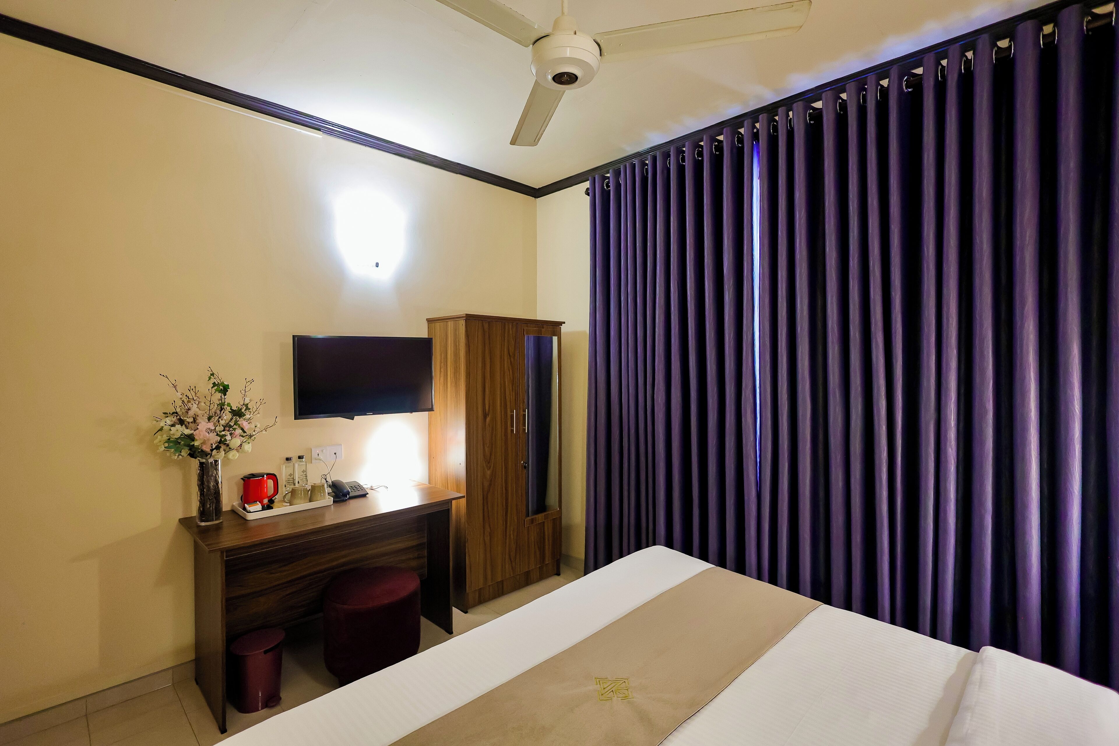 A hotel room with a bed, a desk with a flat-screen TV, a wooden wardrobe, a small red stool, a vase with flowers, and purple curtains.