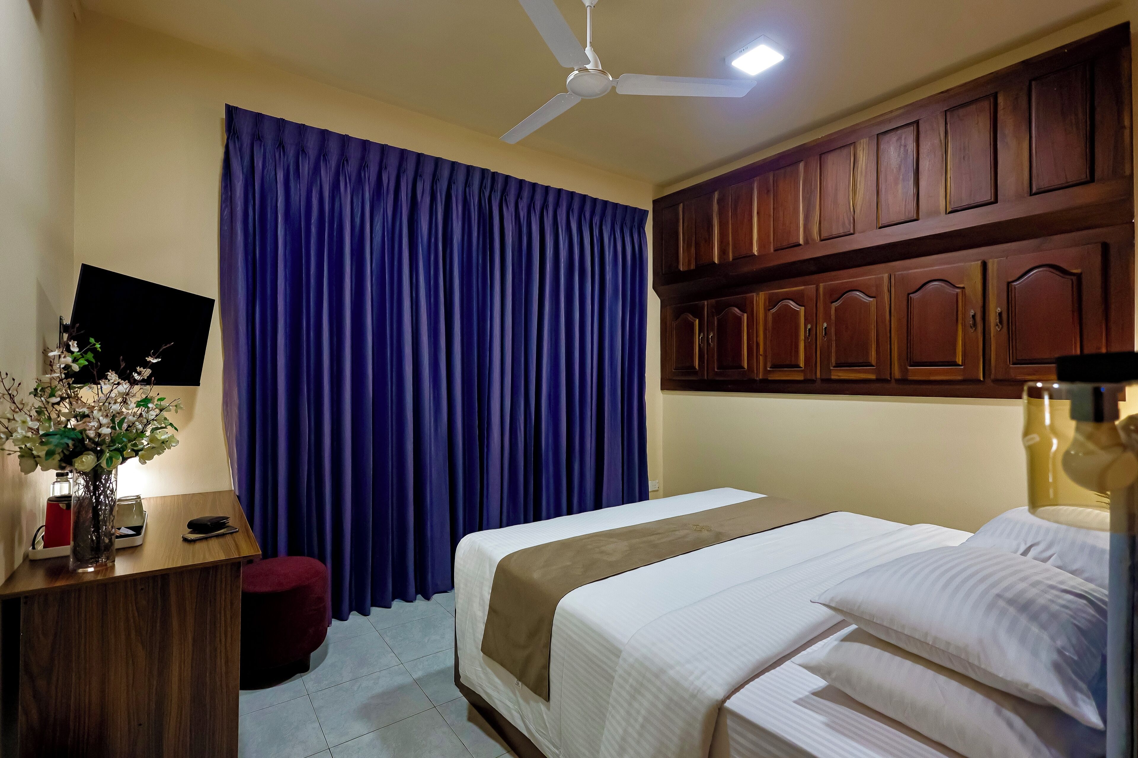 A hotel room with a bed, a desk with a vase of flowers, a ceiling fan, and wooden cabinets.