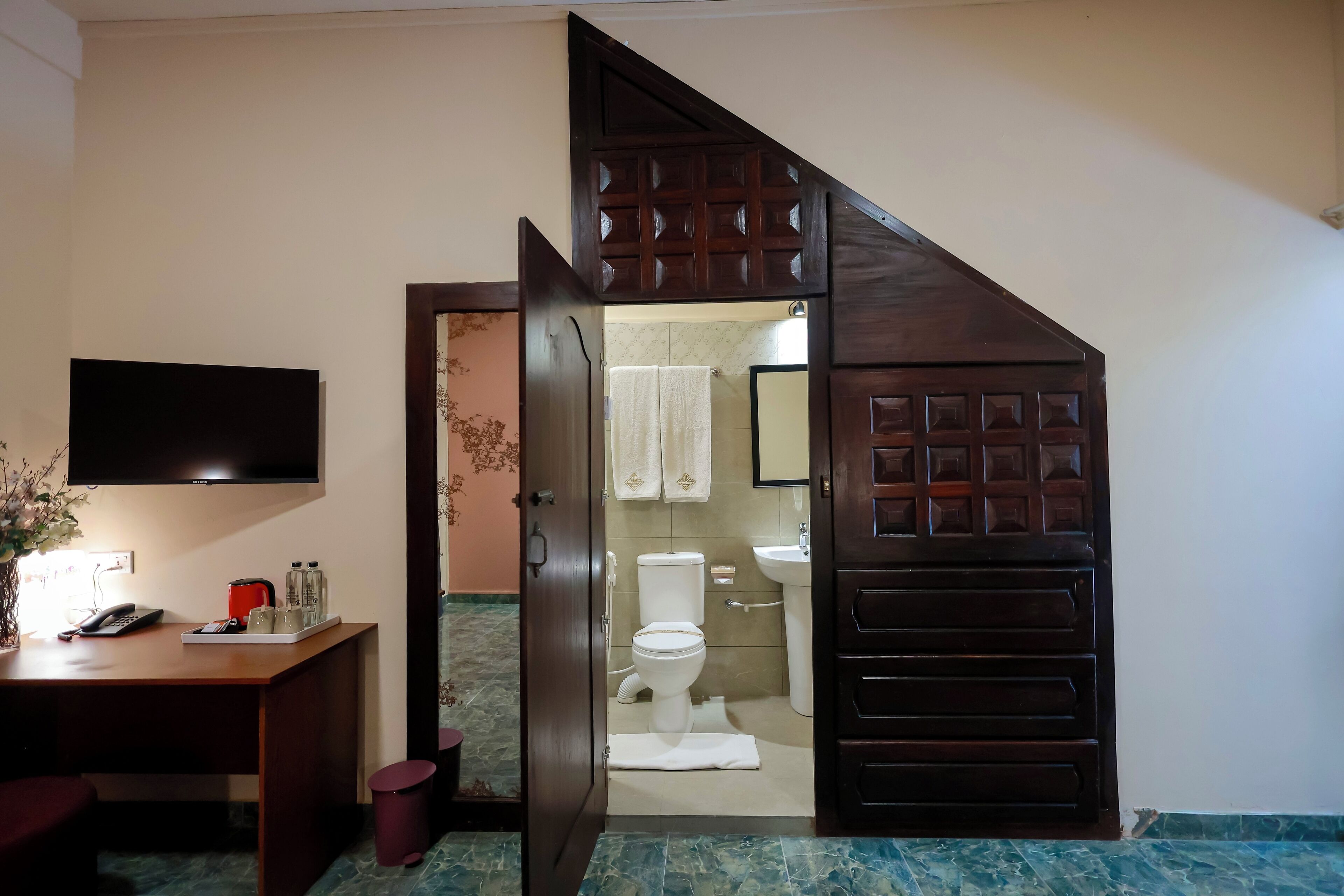 A compact bathroom with a toilet, sink, and mirror. A wooden door leads to a small sitting area with a table and a TV.