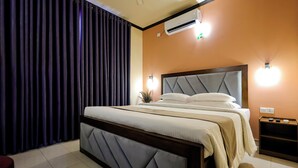 A hotel room with a large bed, dark curtains, an air conditioning unit, and a nightstand with a lamp.