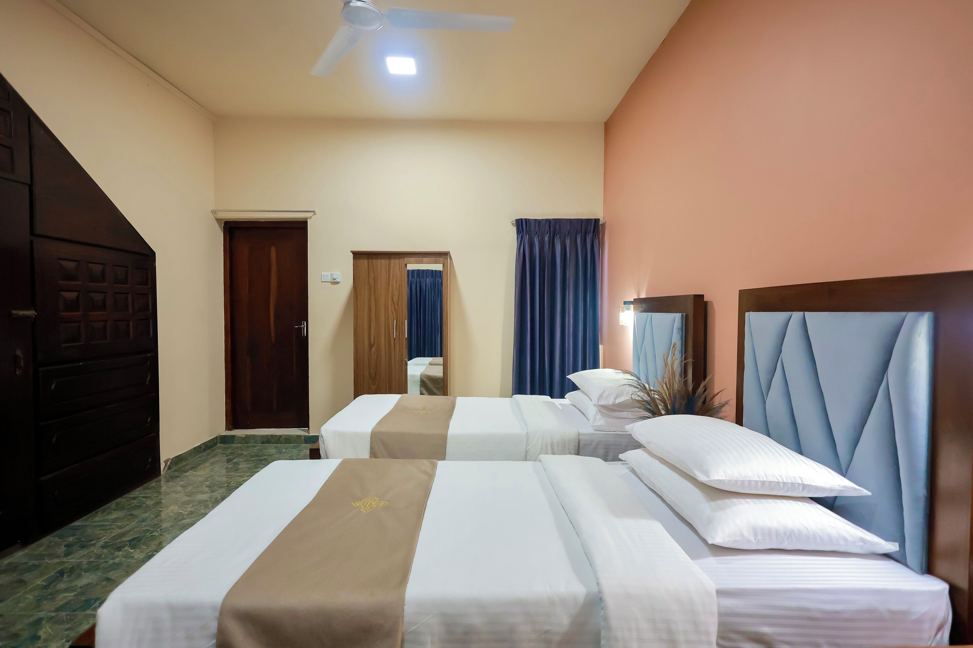 Hotelkamer met twee bedden, een plafondventilator, een donkere houten garderobe en een deur met spiegel en gordijnen.