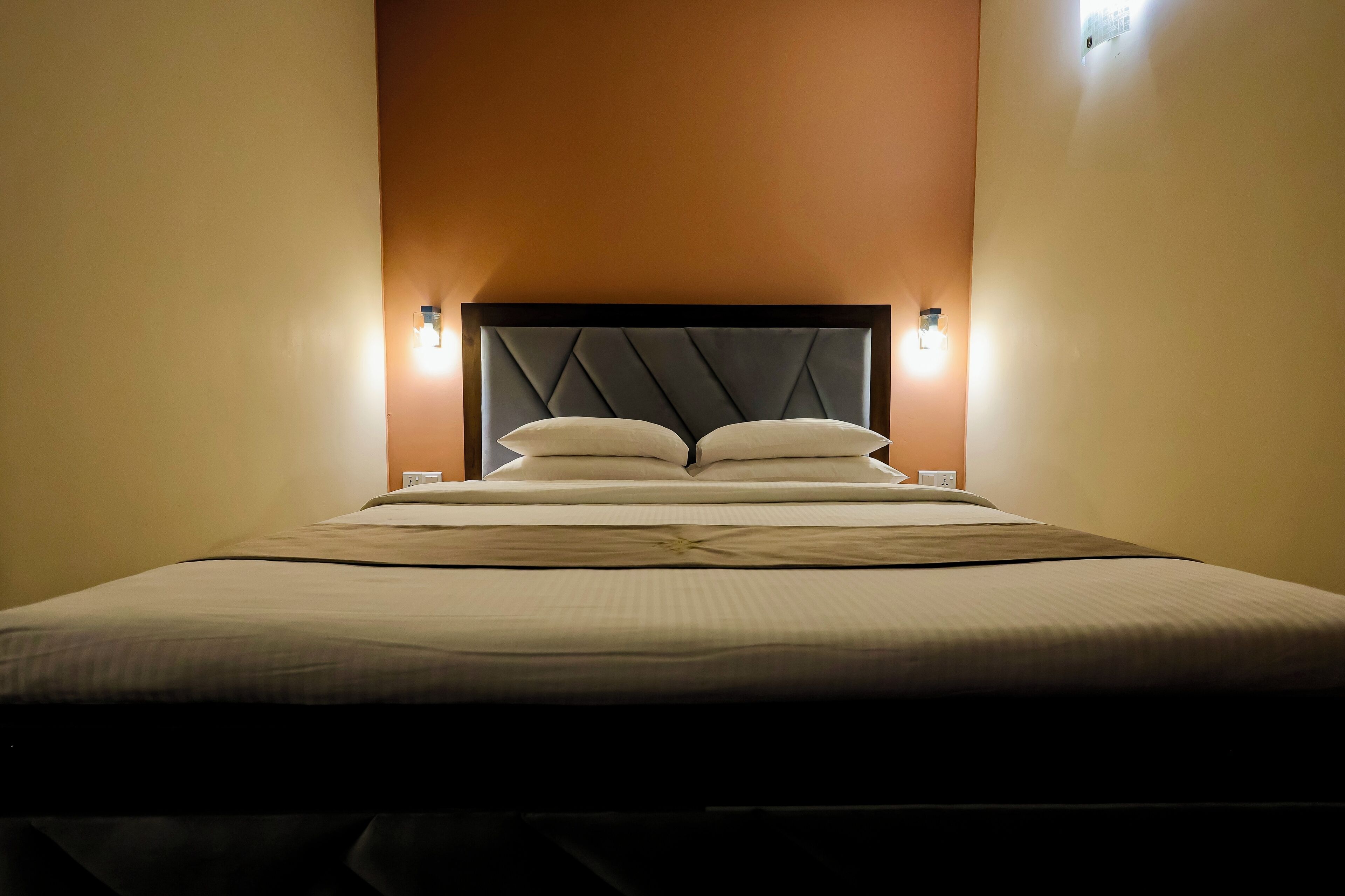 Una cama bien hecha con cabecera, dos lámparas de noche y una lámpara fijada a la pared.