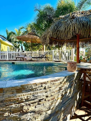 A pool area with a thatched umbrella, lounge chairs, and a white fence.
