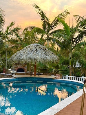 A swimming pool with a thatched-roof structure, surrounded by palm trees and a stone wall.