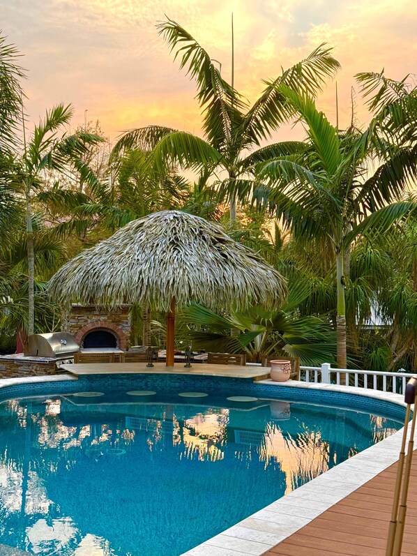 A swimming pool with a thatched-roof structure, surrounded by palm trees and a stone wall.