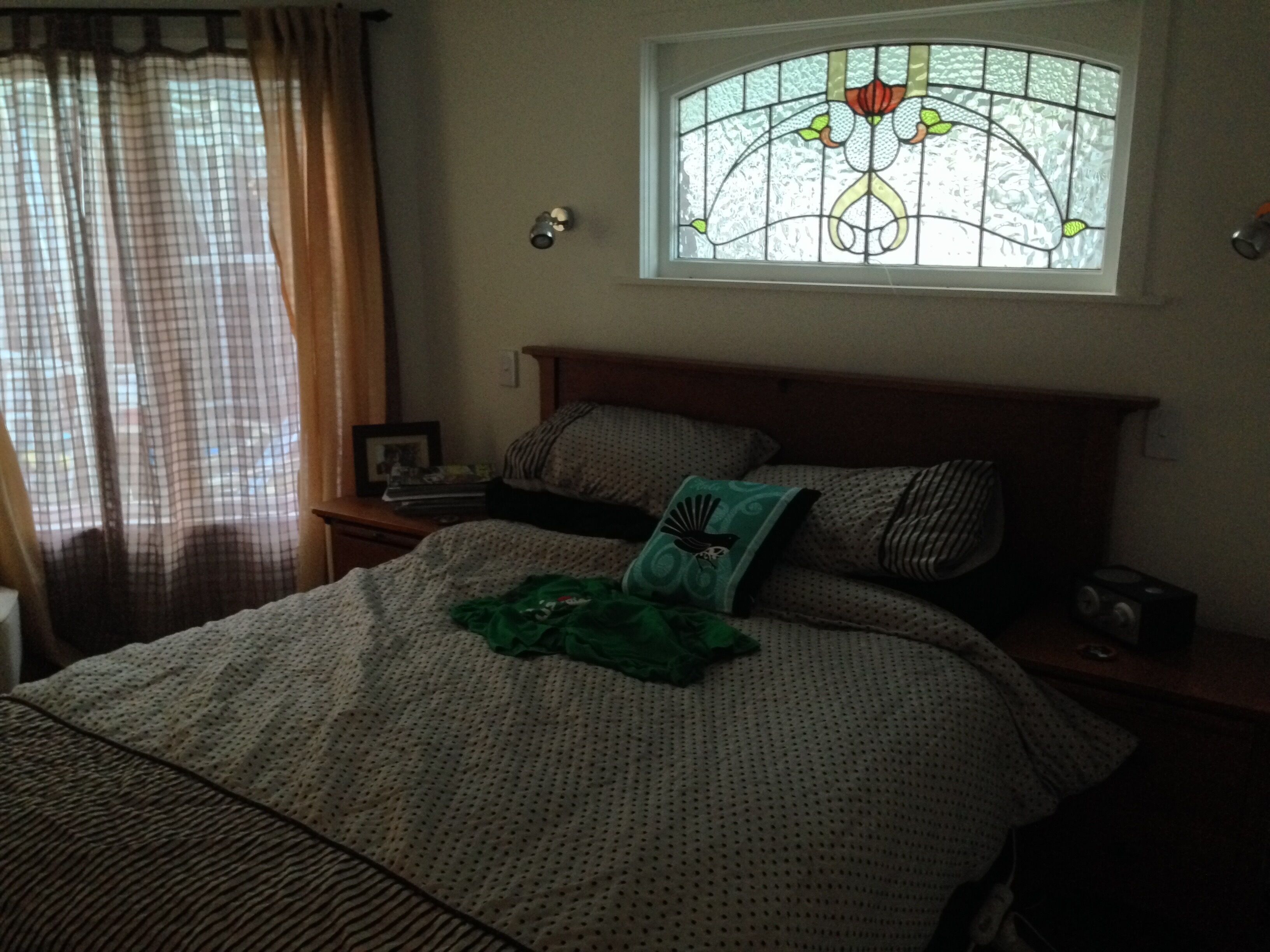 Un dormitorio con cama, mesita de noche y cortina. Ventana con diseño de vitral.