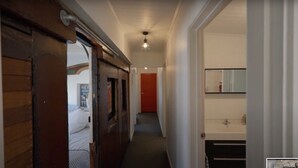 A narrow hallway with a wooden door on the left, a red door at the end, and a bathroom with a mirror and sink on the right.