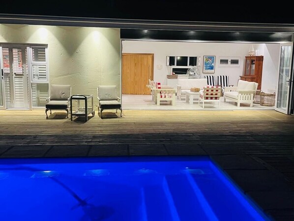 Un área junto a la piscina con un muelle de madera, una piscina con iluminación azul y una zona de estar con muebles blancos.