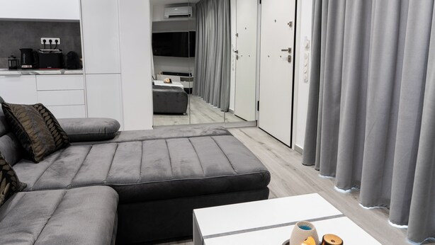 Ein modernes, minimalistisches Wohnzimmer mit einem grauen Ecksofa, einem weißen Couchtisch mit Dekoration und Blick in ein Schlafzimmer mit einem großen Spiegel und grauen Vorhängen.