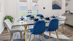 A dining area with a glass table surrounded by blue chairs, a white sofa, and a painting on the wall. - Spacious Yard+Pool Table at 5BR Modern Getaway (North Las Vegas)