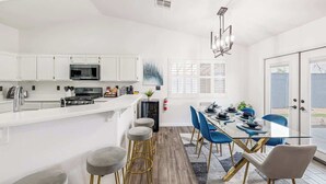 A modern kitchen with white cabinets, a central island with stools, and a dining area with blue chairs and a glass table. - Spacious Yard+Pool Table at 5BR Modern Getaway (North Las Vegas)
