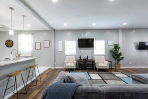 A modern living room with a grey sofa, wooden bar stools, a flat-screen TV, and a dining area with striped chairs.