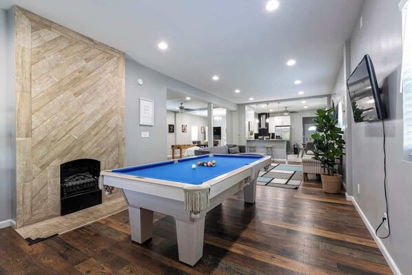 A modern living room with a pool table, a fireplace, and a kitchen in the background.