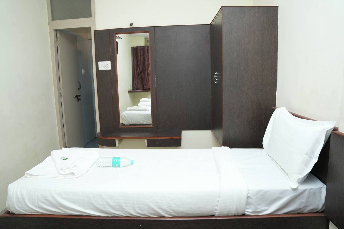A single bed with white bedding, a wooden wardrobe, a mirror, and a small table with a bottle.