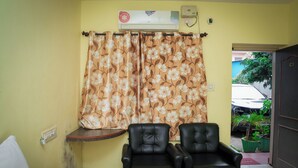 A room with a yellow wall, a floral curtain, two black leather chairs, a small table, and an air conditioning unit. - The Vishnu Residency (Chikkamagaluru)