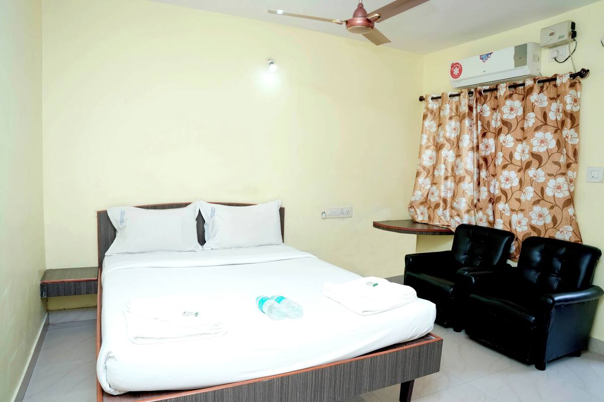 A hotel room with a bed, a ceiling fan, an air conditioning unit, a small table, and two black leather chairs.