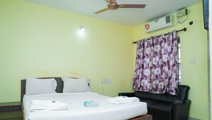 A hotel room with a bed, a ceiling fan, an air conditioning unit, a black leather sofa, and a window with floral curtains. - The Vishnu Residency (Chikkamagaluru)