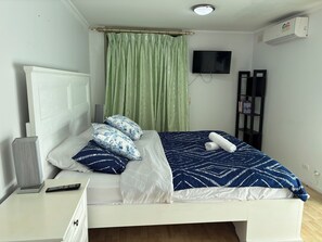 A neatly made bed with patterned bedding, a bedside table with a lamp and remote, a TV on the wall, and a bookshelf. - Sunflower Homes Classy and Cozy (Para Hills)