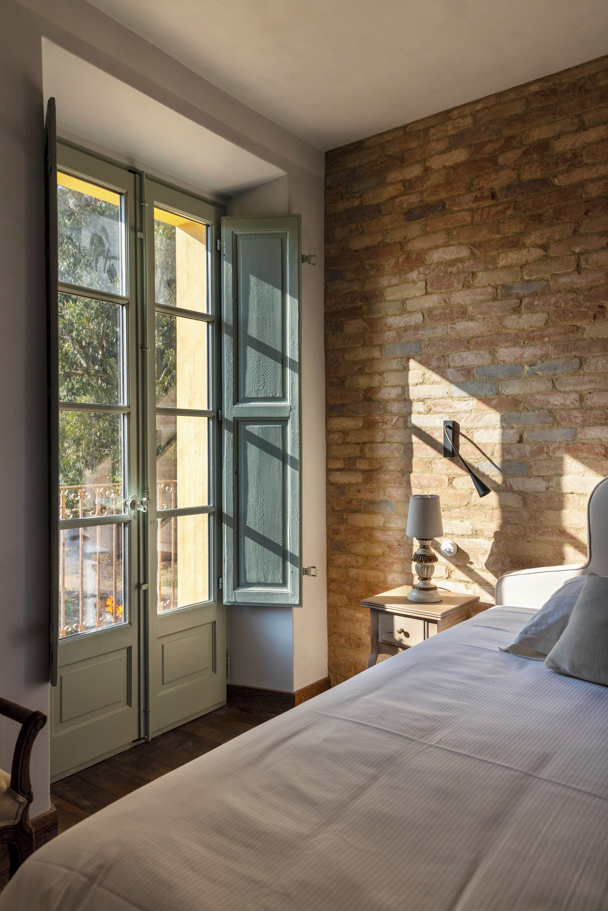 A bedroom with a large bed, a nightstand with a lamp, a brick wall, and a window with open shutters.