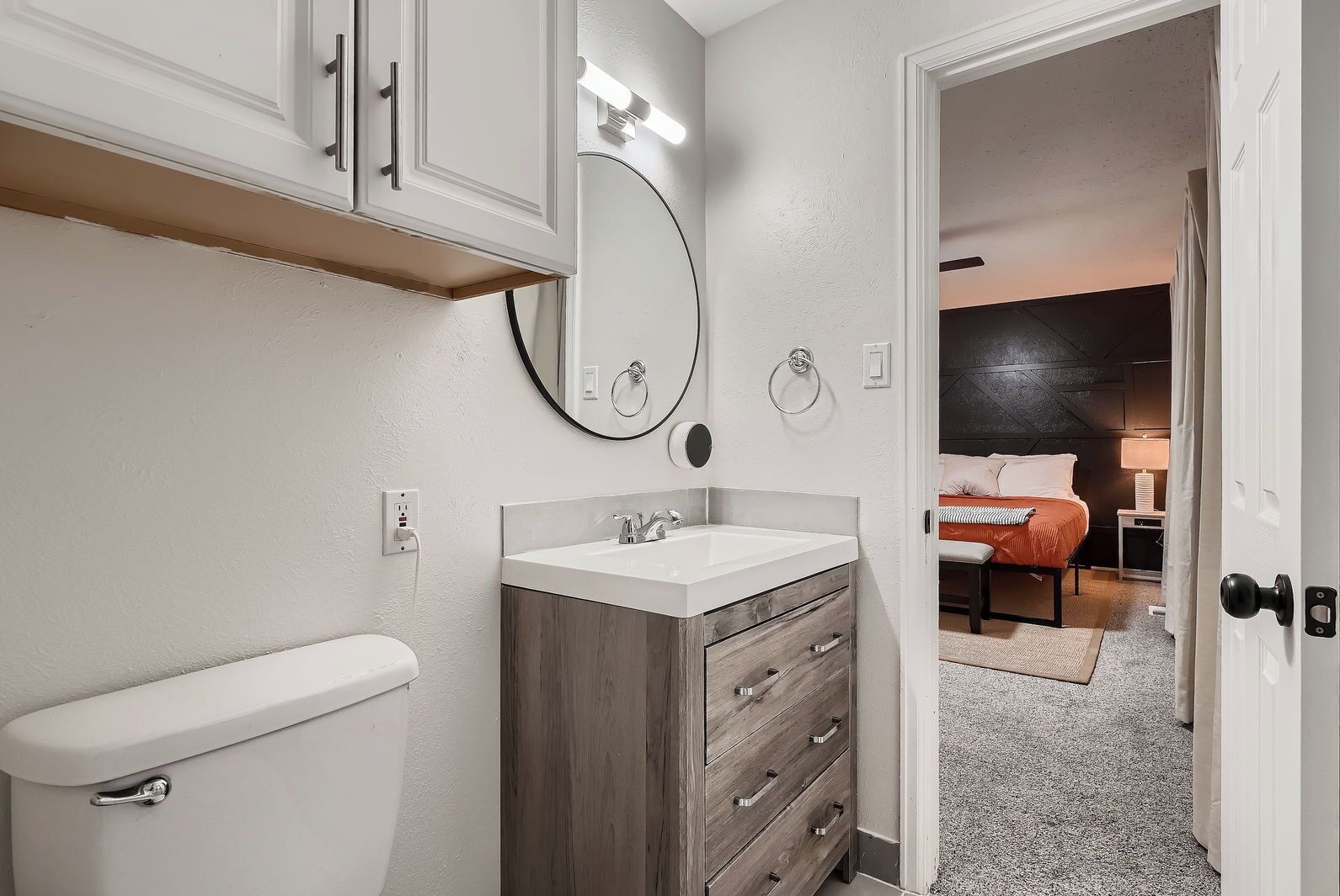 Un bagno moderno con il water, un lavabo e uno specchio. Si intravede una camera da letto sullo sfondo, con un letto e un comodino.