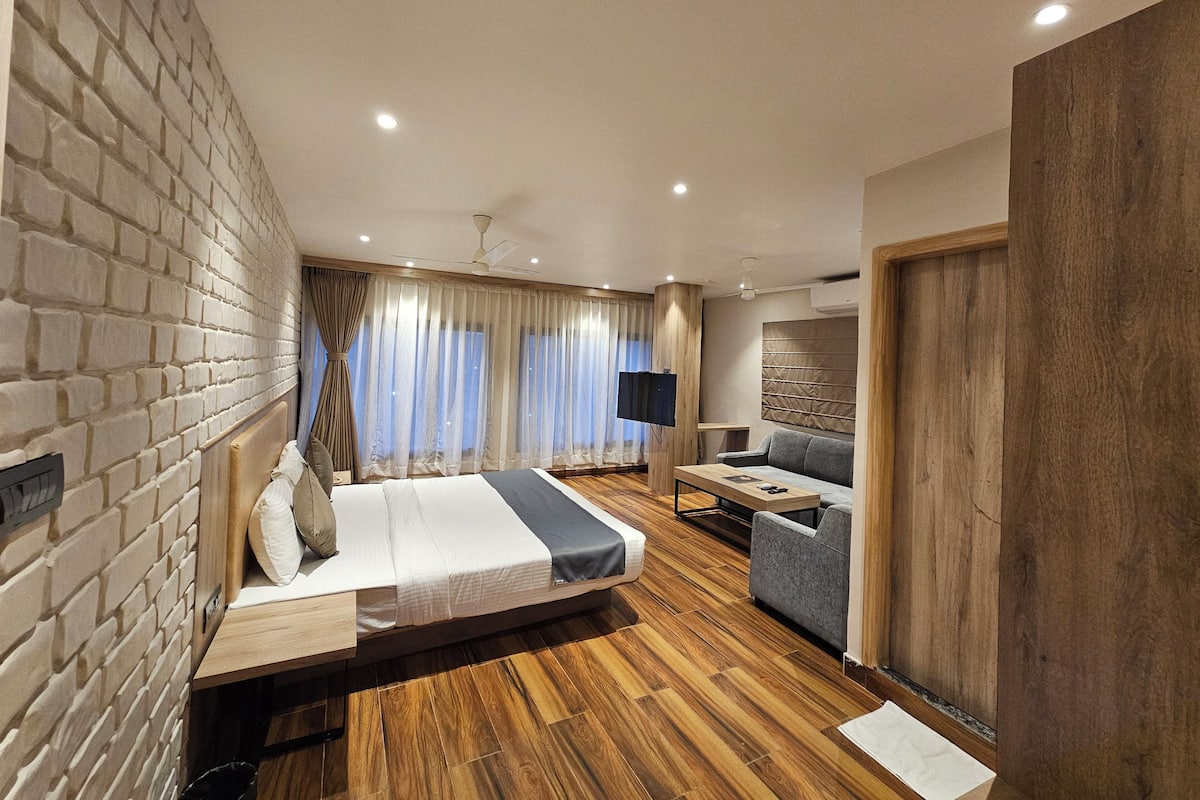 A modern hotel room with a brick-patterned wall, a wooden floor, a bed with a grey and white duvet, a grey sofa, a small table, and a TV.