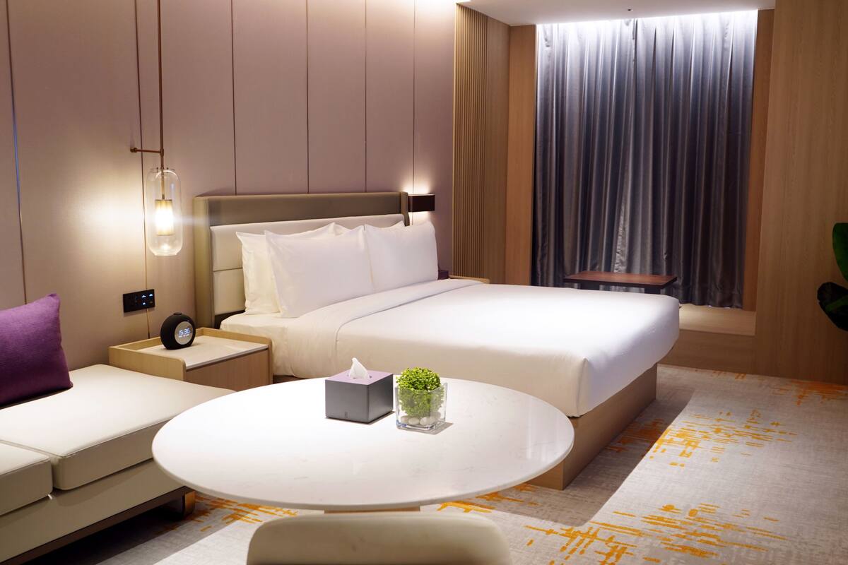 Quarto de hotel moderno com uma cama grande, mesas de cabeceira, uma mesa de centro redonda e um vaso de planta.