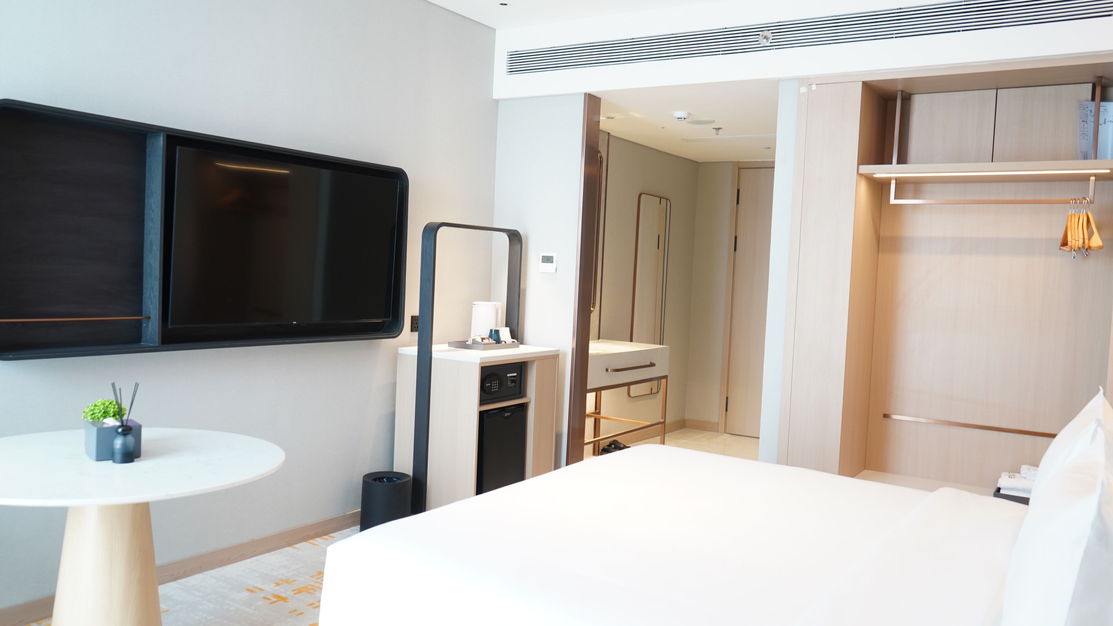 A modern hotel room with a large bed, a round table with a plant, a TV mounted on the wall, a wardrobe, and a bathroom visible through an open door.
