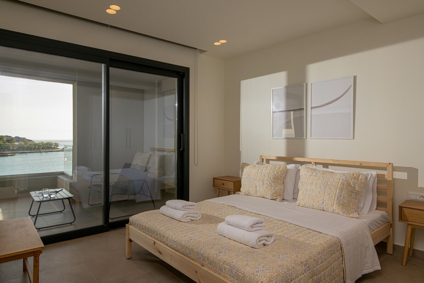 A bedroom with a bed, bedside tables, a small table, and a chair. There is a sliding glass door leading to an outdoor area with a view of the sea.