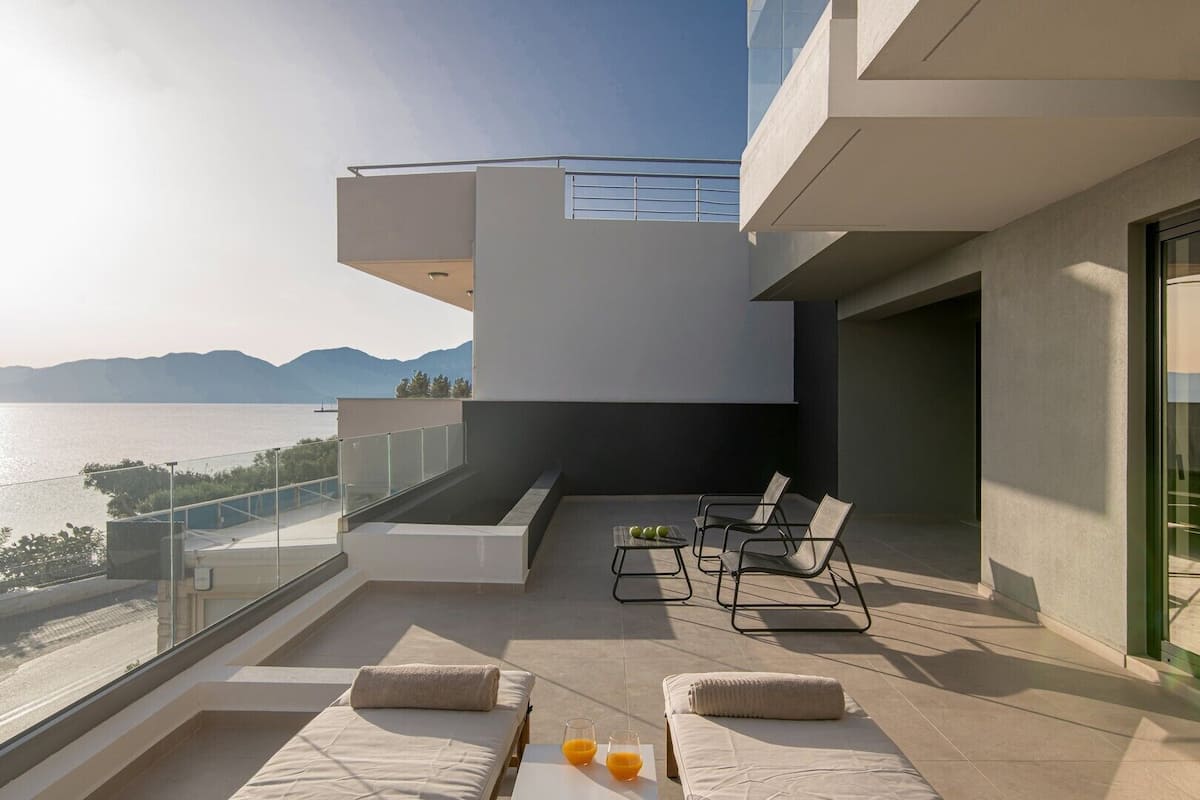 Un balcon moderne doté d’une piscine, de chaises longues et d’une balustrade en verre, offrant une vue sur un plan d’eau et des montagnes.
