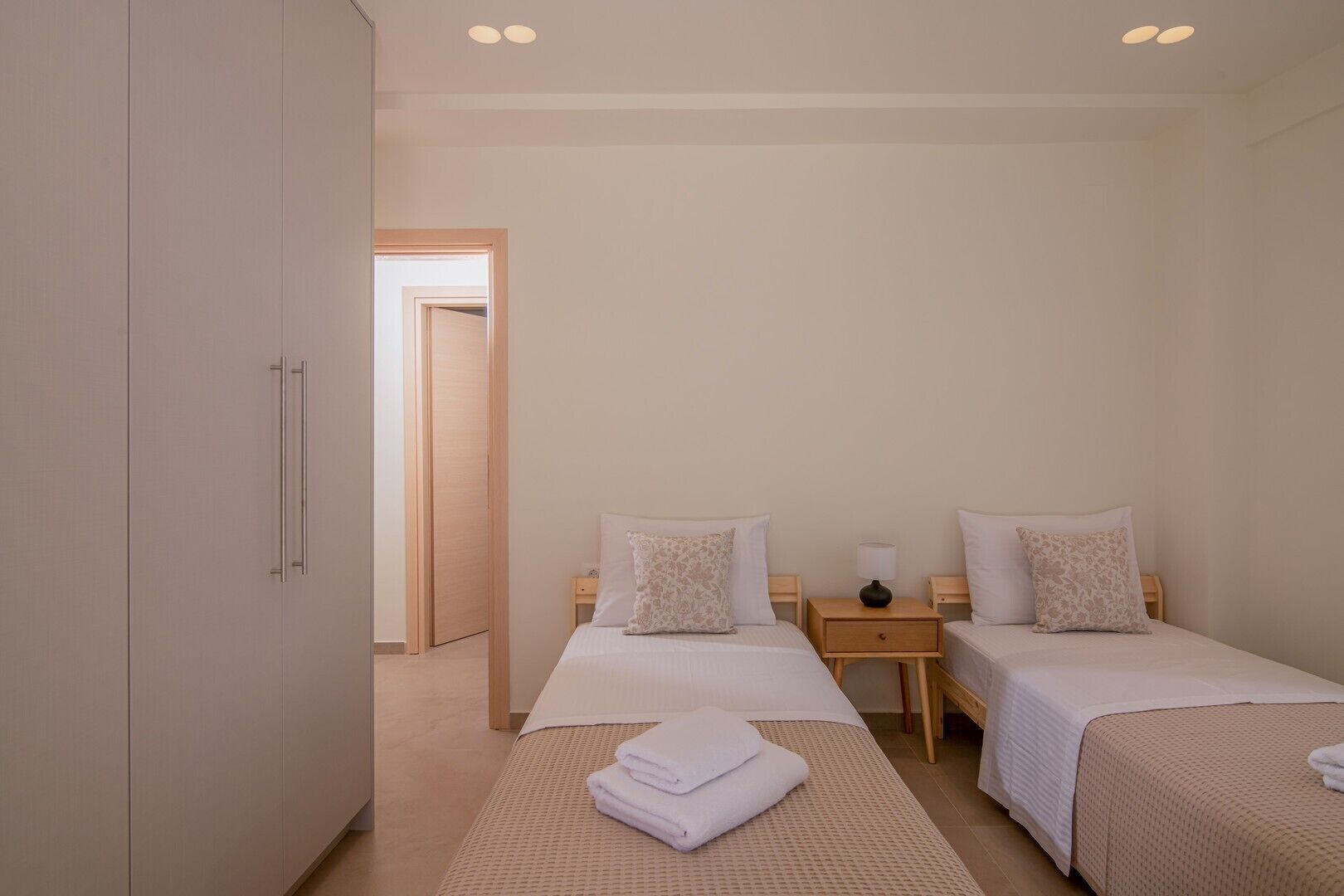 Une chambre d’hôtel avec deux lits simples, une armoire, une petite table de chevet avec une lampe et une porte donnant sur une autre pièce.