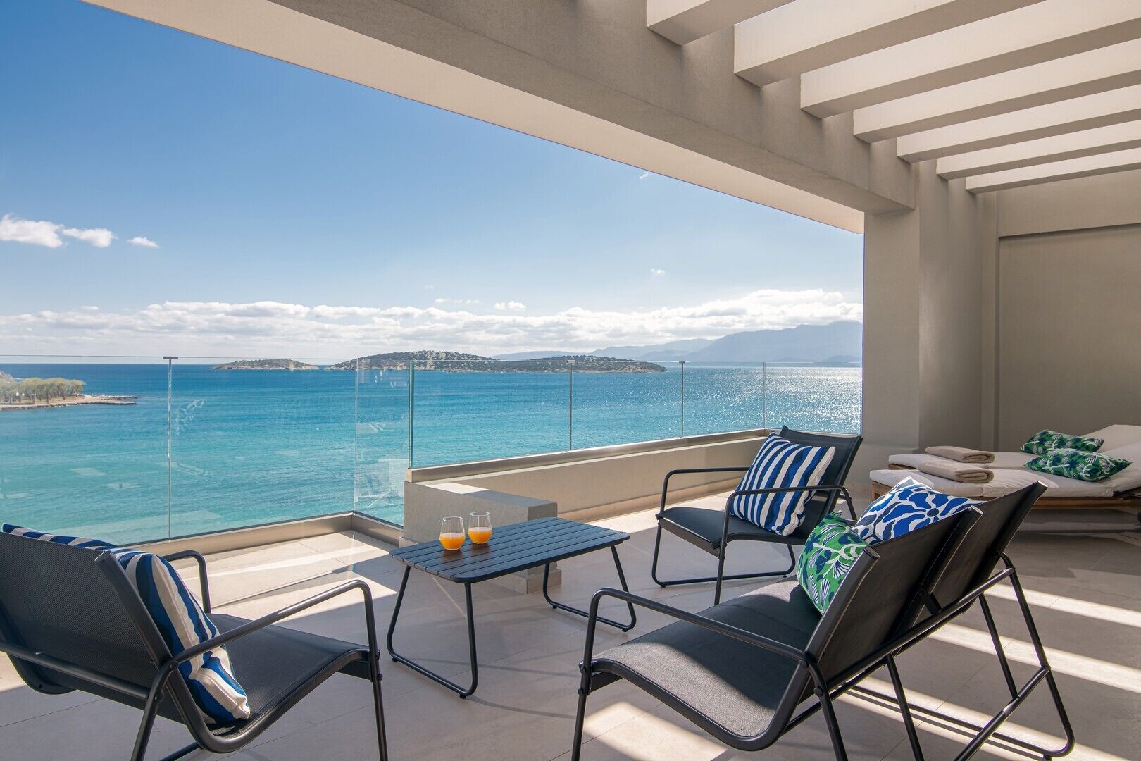 A balcony with a clear view of the sea, two chairs, a small table, and a sofa with cushions.